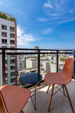 Un balcón con dos sillas y una mesita, con vista a un paisaje urbano que incluye edificios y cielo despejado.