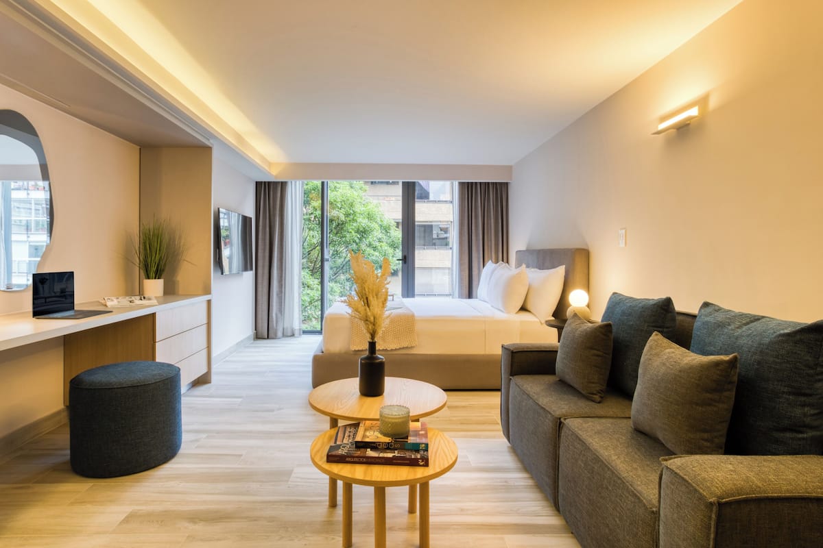 A modern hotel room with a sofa, a desk, and a bed. There is a window with a view of buildings and trees.