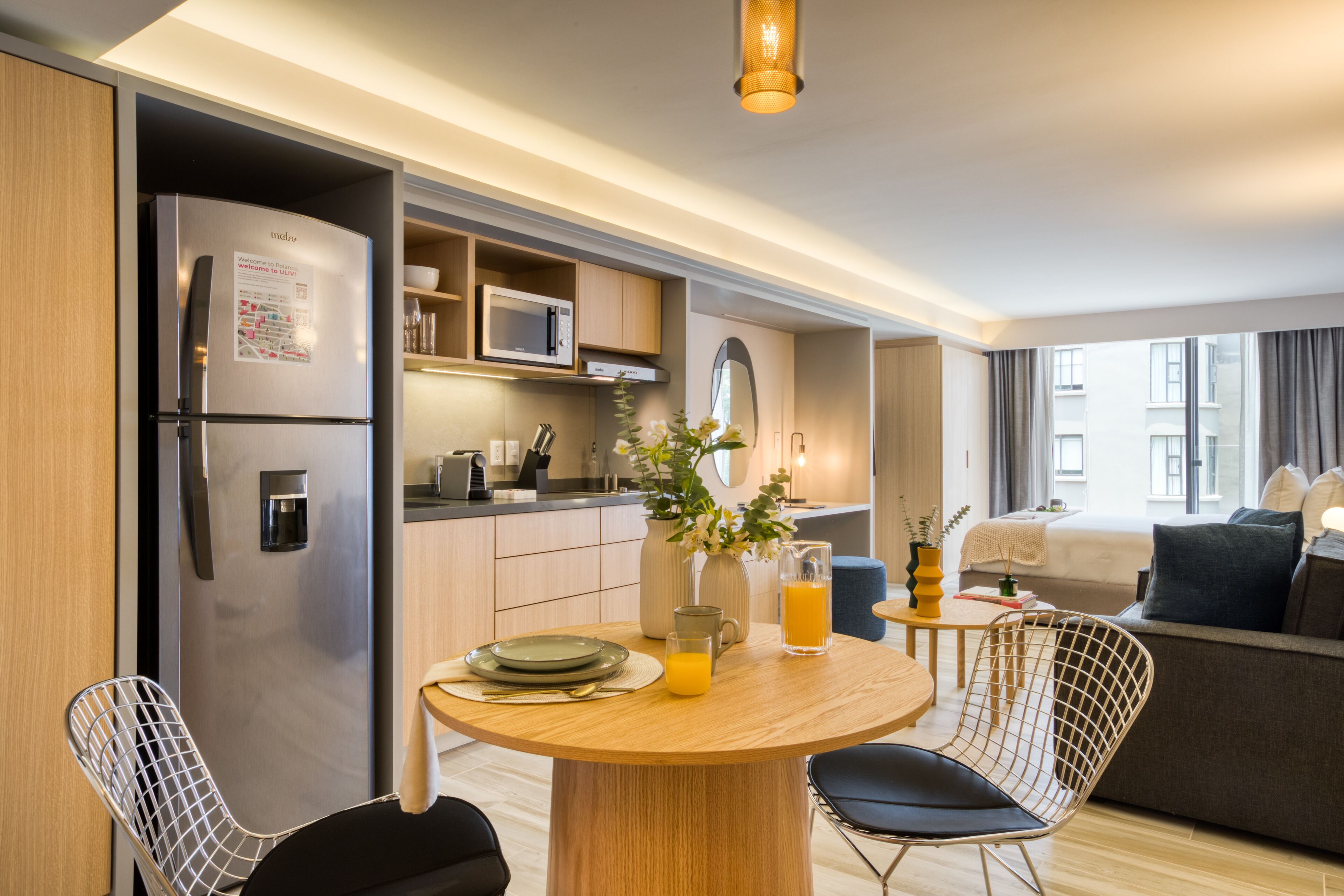 Eine moderne Küche mit integriertem Kühlschrank, Mikrowelle und Kaffeemaschine. Es gibt einen Essbereich mit einem Holztisch, Stühlen und einer Vase mit Blumen. Im Hintergrund ist ein Bett mit Blick auf die Stadt zu sehen.
