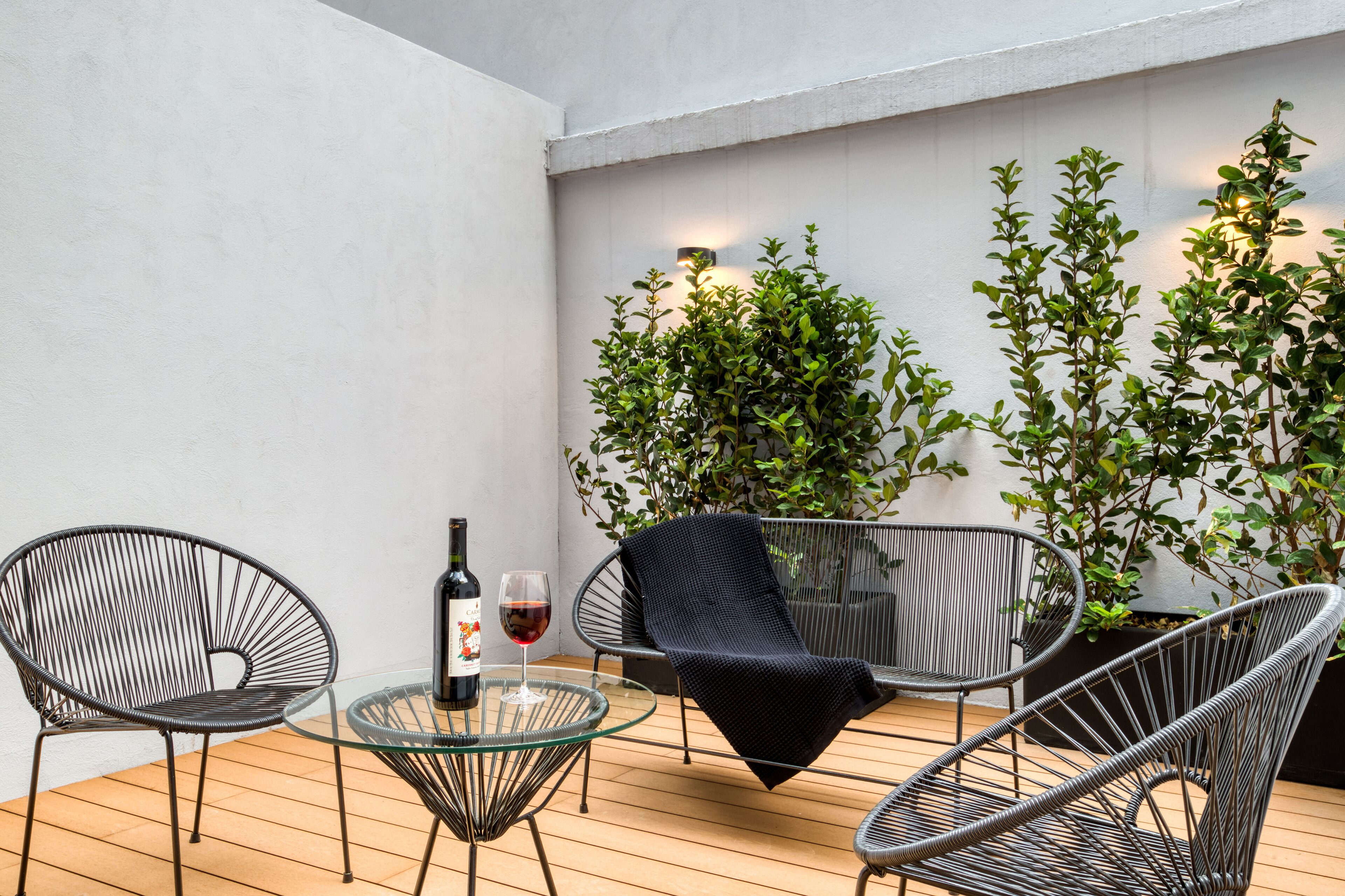 Un espacio al aire libre moderno con sillas de metal y una mesa de vidrio, una botella de vino tinto, y plantas en macetas frente a una pared blanca.