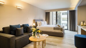 A modern hotel room with a sofa, a round wooden table with a vase of flowers, a bed with a large window, and a desk area. - ULIV Parque Lincoln (Mexico City)