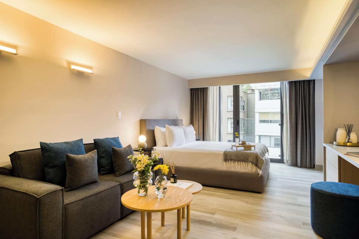 A modern hotel room with a sofa, a round wooden table with a vase of flowers, a bed with a large window, and a desk area.