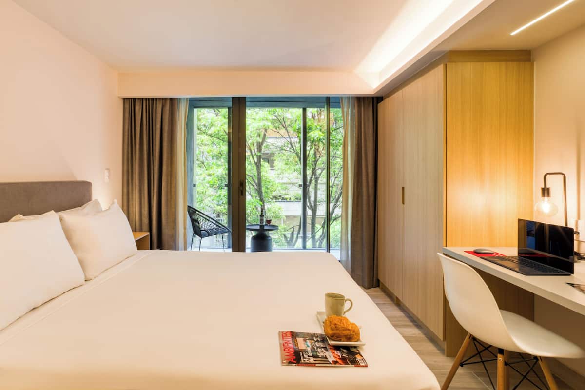A hotel room with a large bed, a desk with a laptop, a chair, and a view of greenery outside.