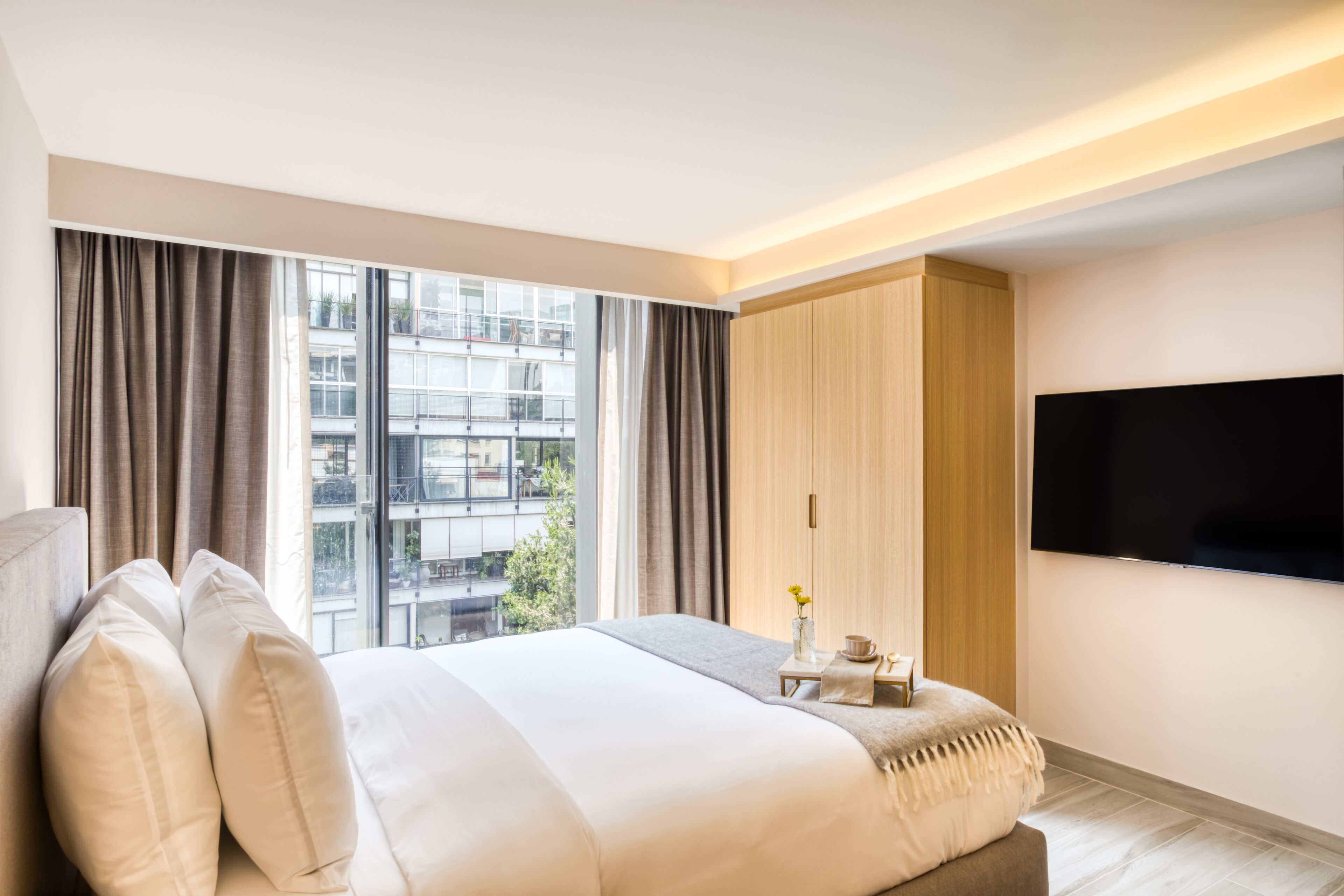 A modern hotel room with a large bed, a flat-screen TV, and a view of the city through large windows.