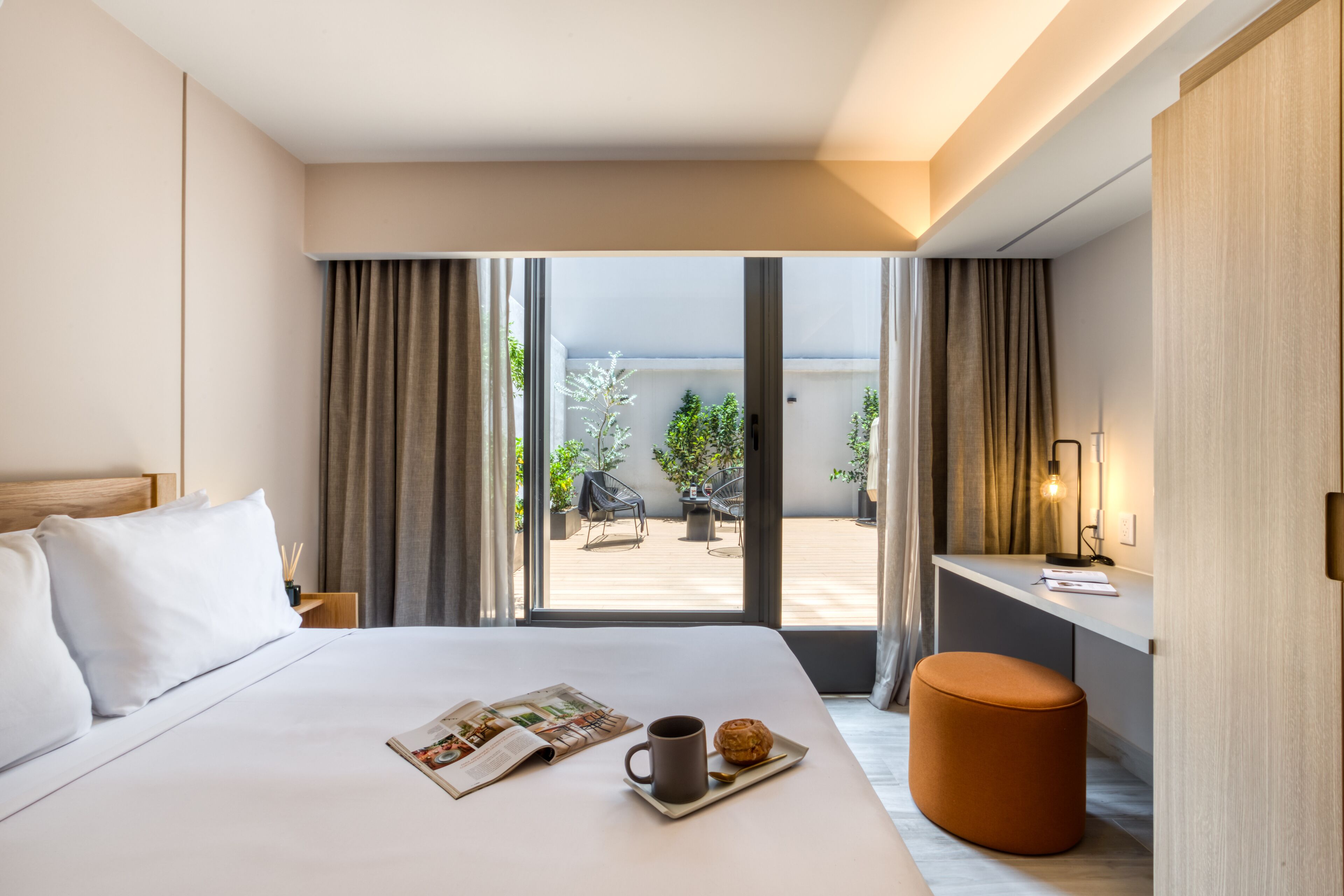 Una habitación de hotel con una cama, una bandeja de desayuno con un muffin y café, un escritorio con una lámpara, y vistas del patio exterior.