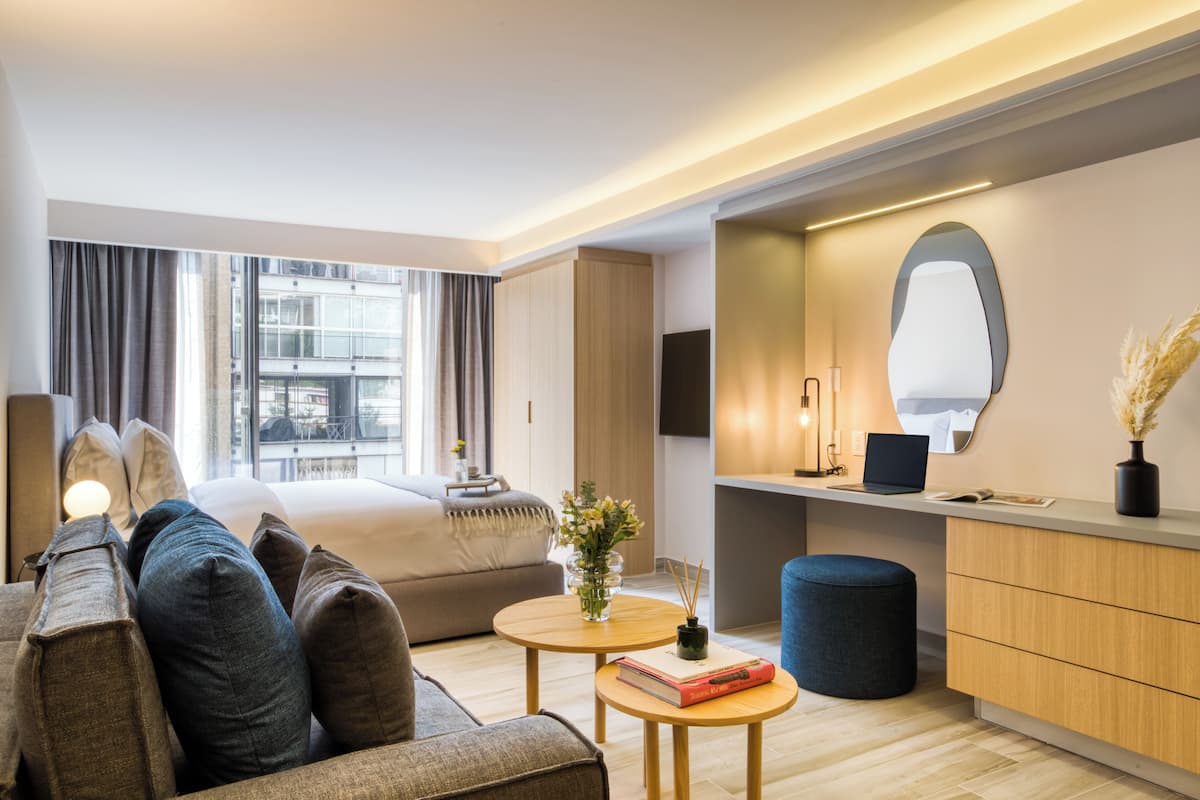 A modern hotel room with a bed, a sofa, a small round table with a book, a mirror, and a desk area with a laptop.