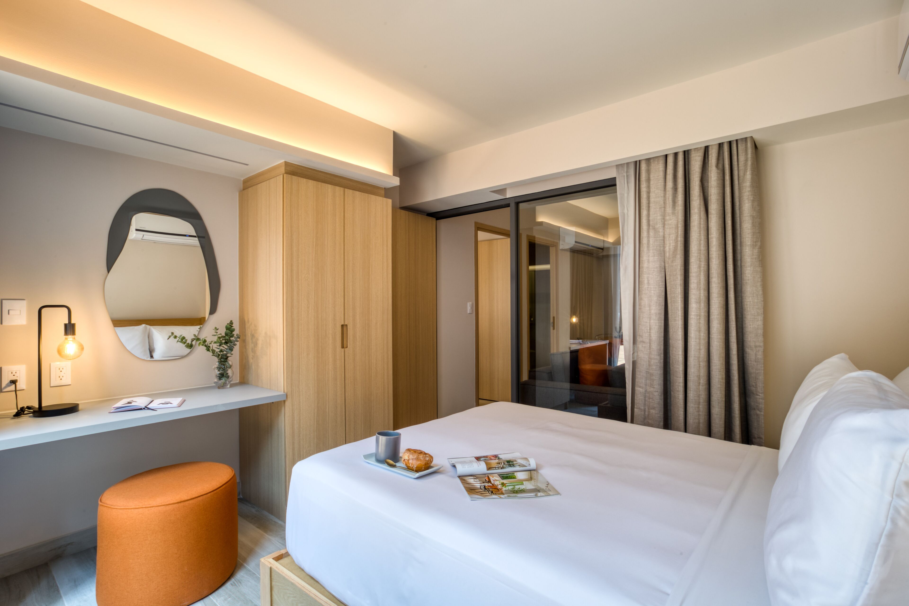A modern hotel room with a neatly made bed, a small desk with a lamp, a mirror, and a large window with curtains.