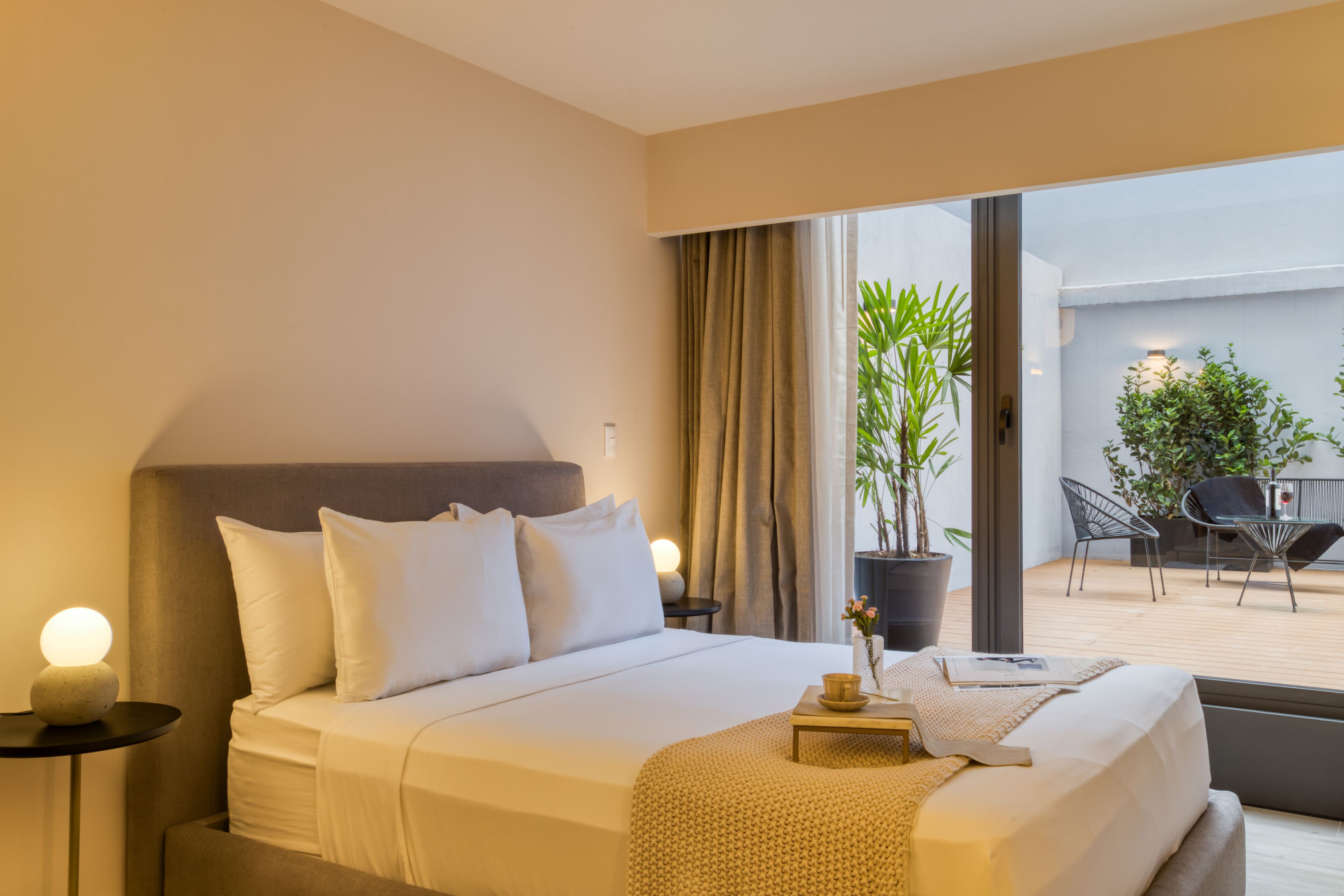 Habitación de hotel con cama, mesita de noche, lámpara y vistas al patio exterior.