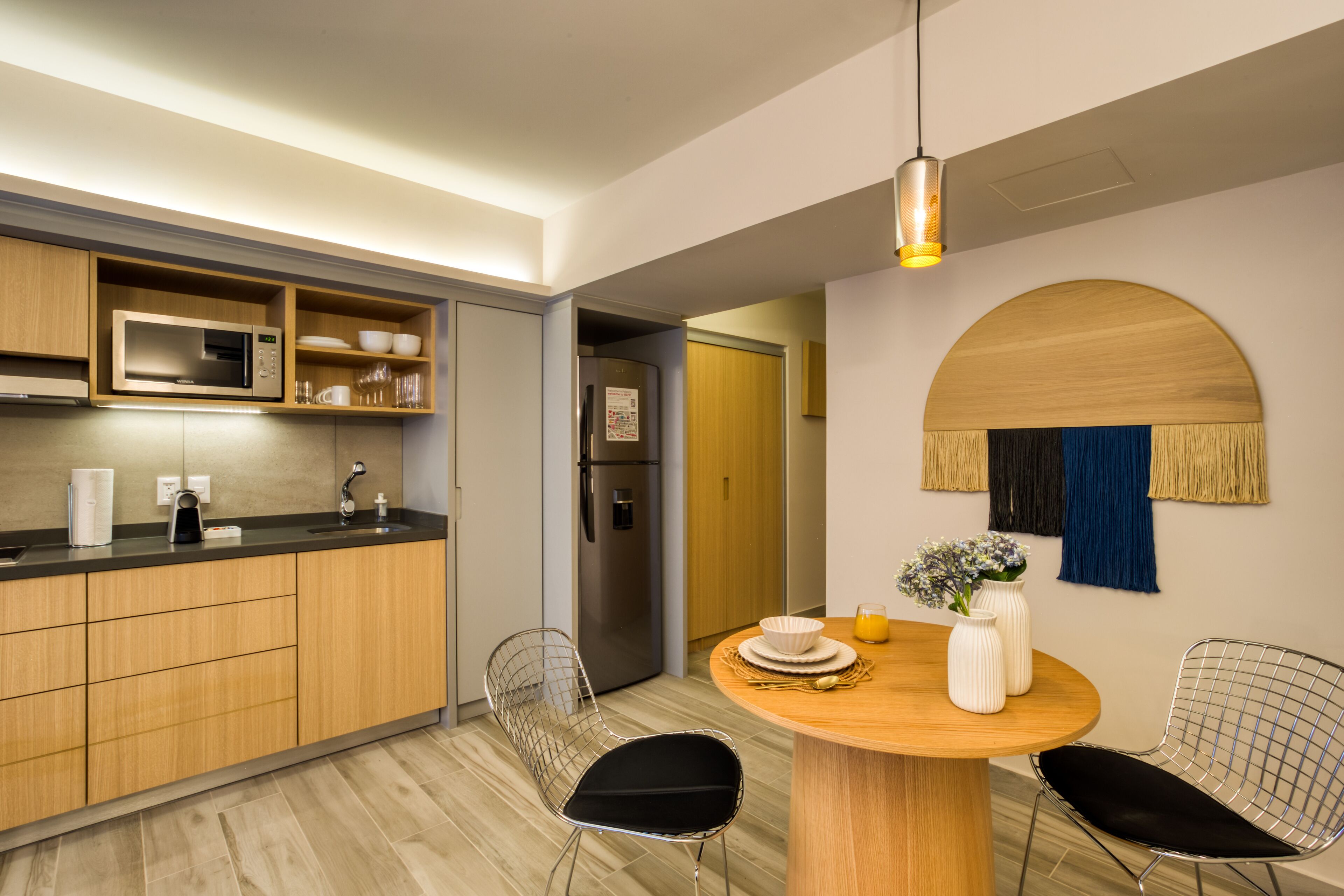A modern kitchen with wooden cabinets, a built-in microwave, and a stainless steel refrigerator. There is a dining area with a round wooden table, two wireframe chairs, and a vase with flowers.