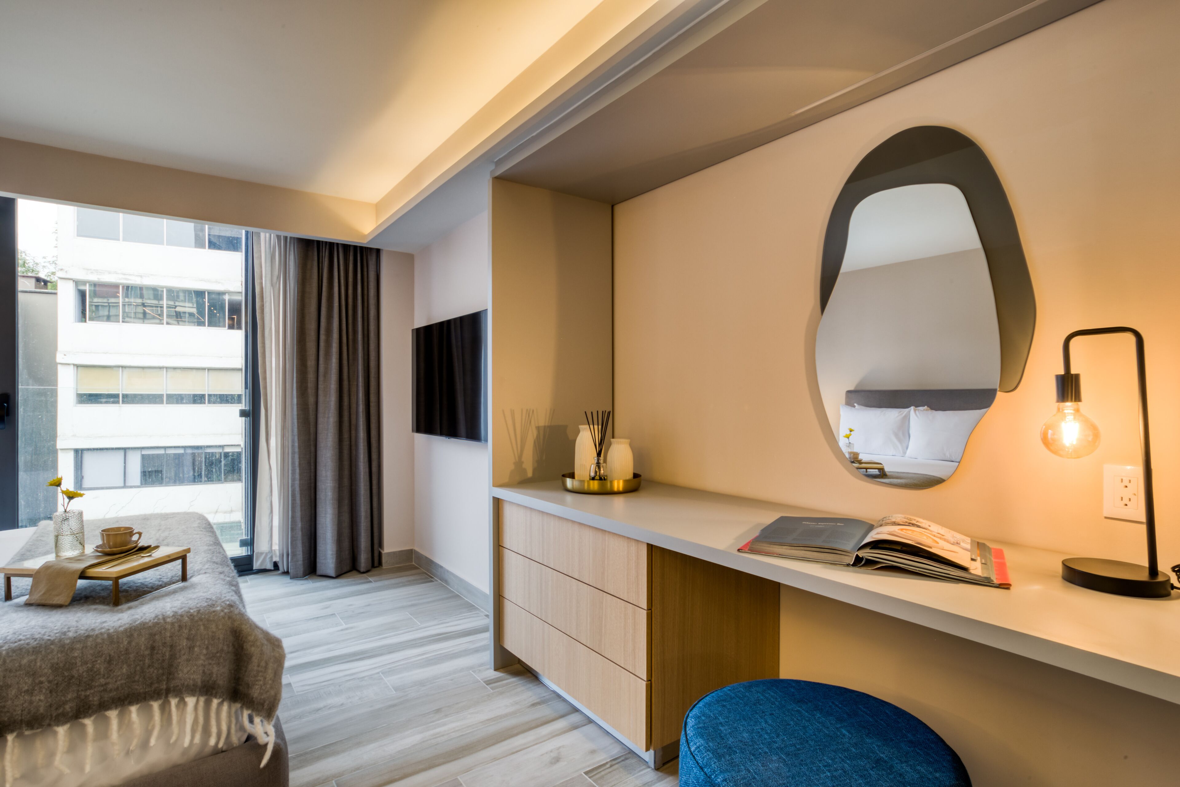Una habitación de hotel moderna con cama, mesita de noche y escritorio con espejo y lámpara.