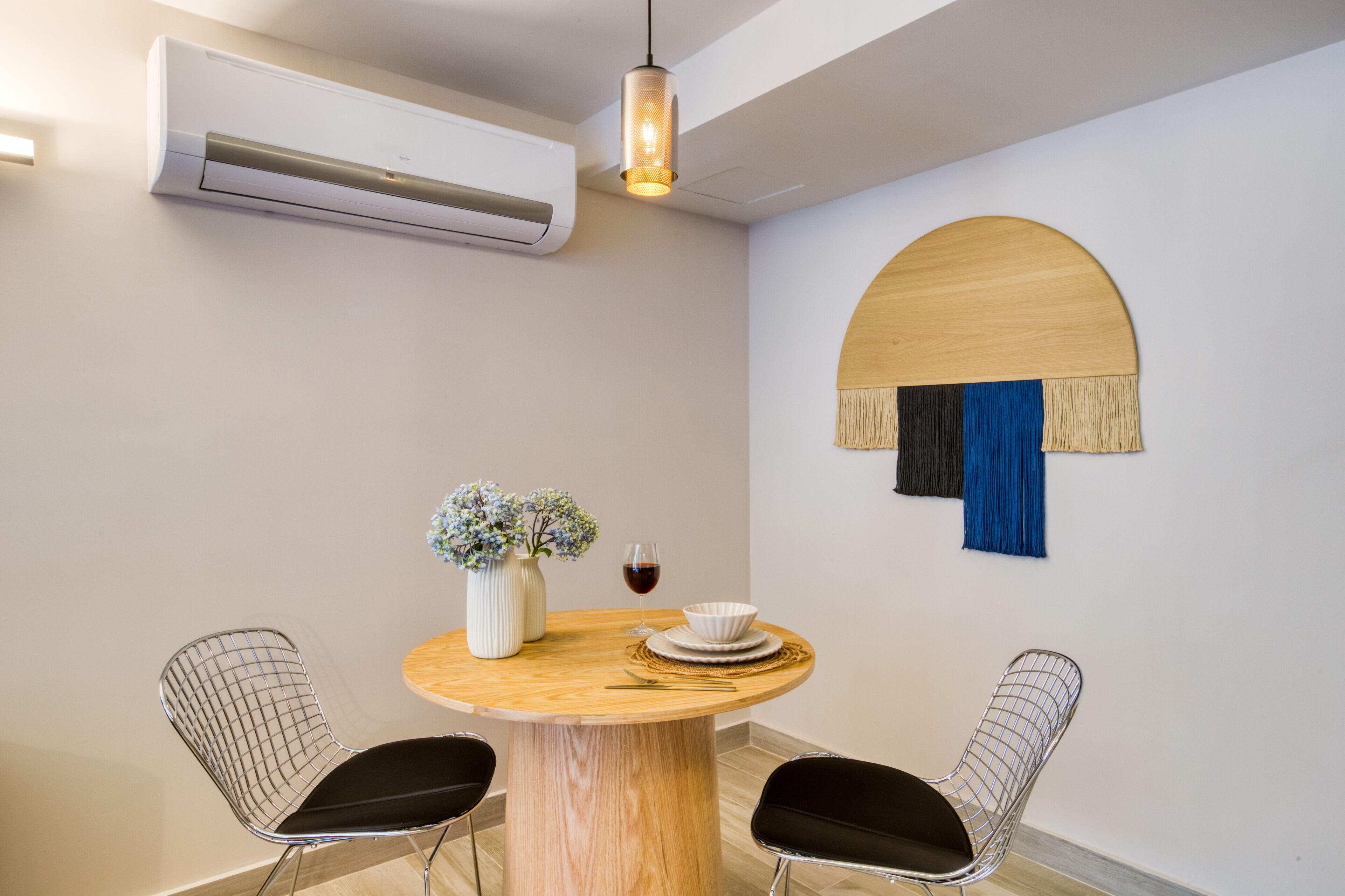 Een eetruimte met een ronde houten tafel, twee open stoelen, een vaas met bloemen, een wijnglas en een wandairconditioner.