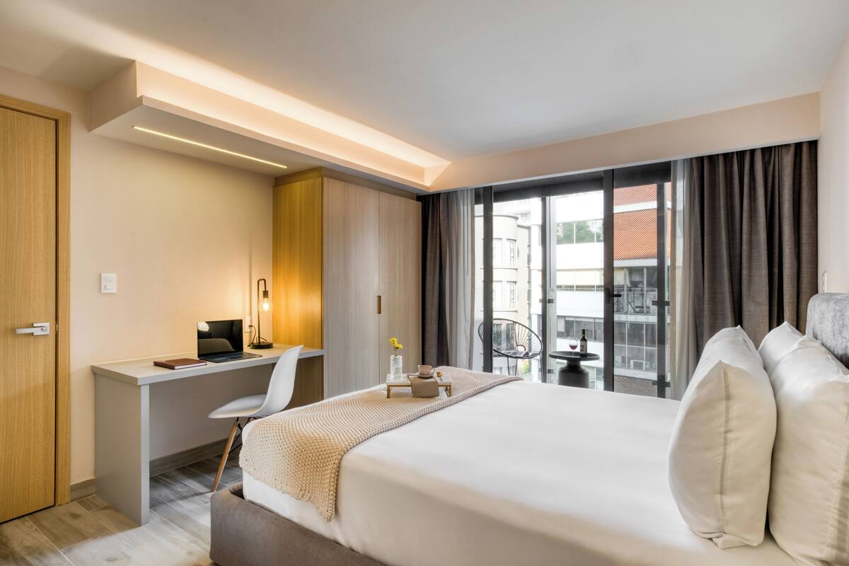 A hotel room with a bed, desk, chair, and a view of the city through large windows.