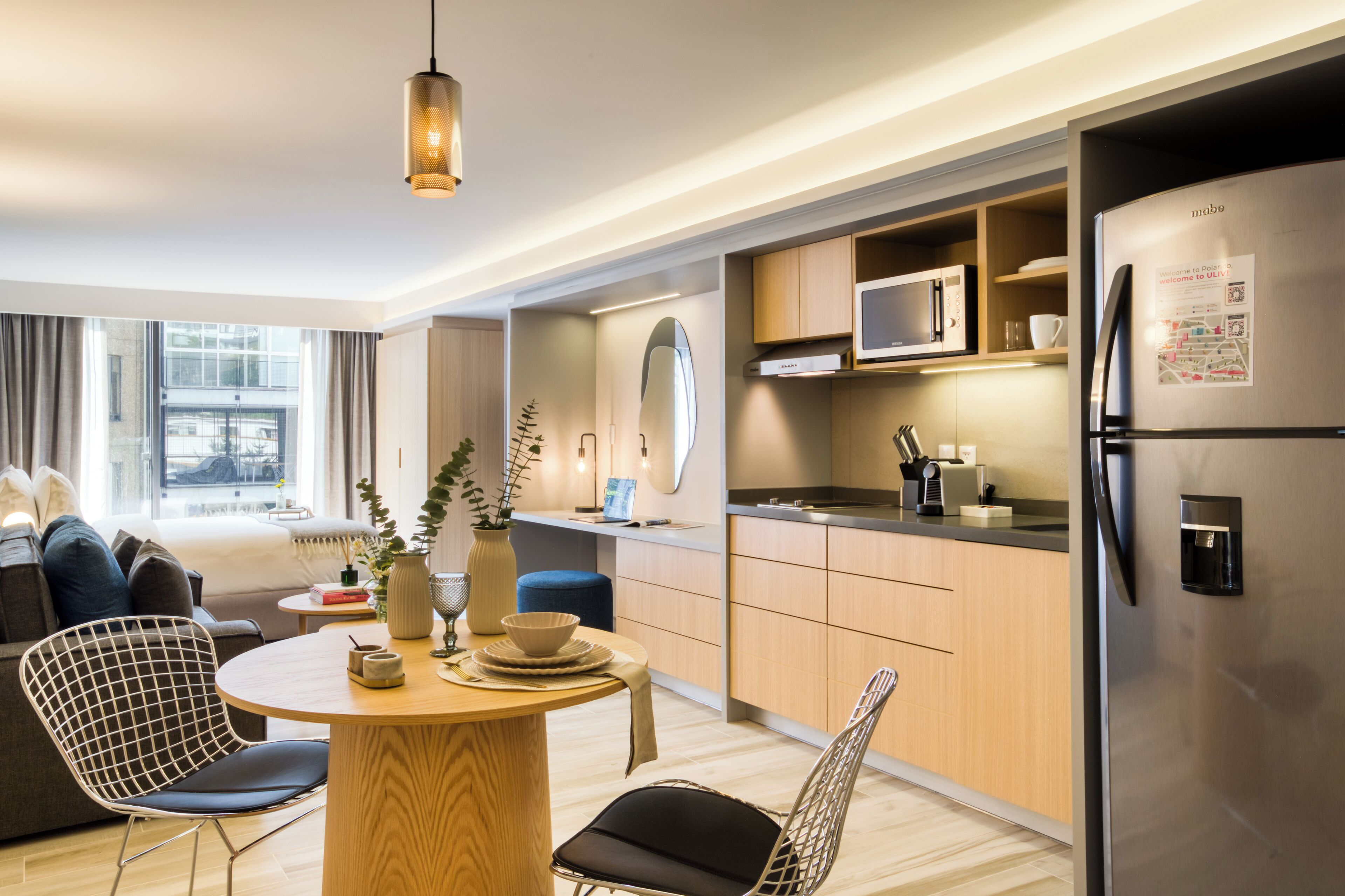 A modern kitchen with a built-in microwave, stainless steel refrigerator, and wooden cabinets. There is a dining area with a round table and chairs.