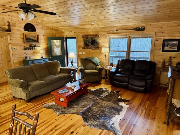 L’intérieur d’un chalet accueillant, avec un plafond en bois, un canapé, deux fauteuils, une table basse et une grande fenêtre.