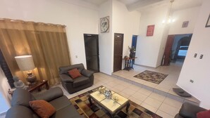 Un salón con sofá, sillón, mesa de centro y alfombra decorativa. Se observa una puerta, un reloj en la pared y una pequeña mesa con lámpara.