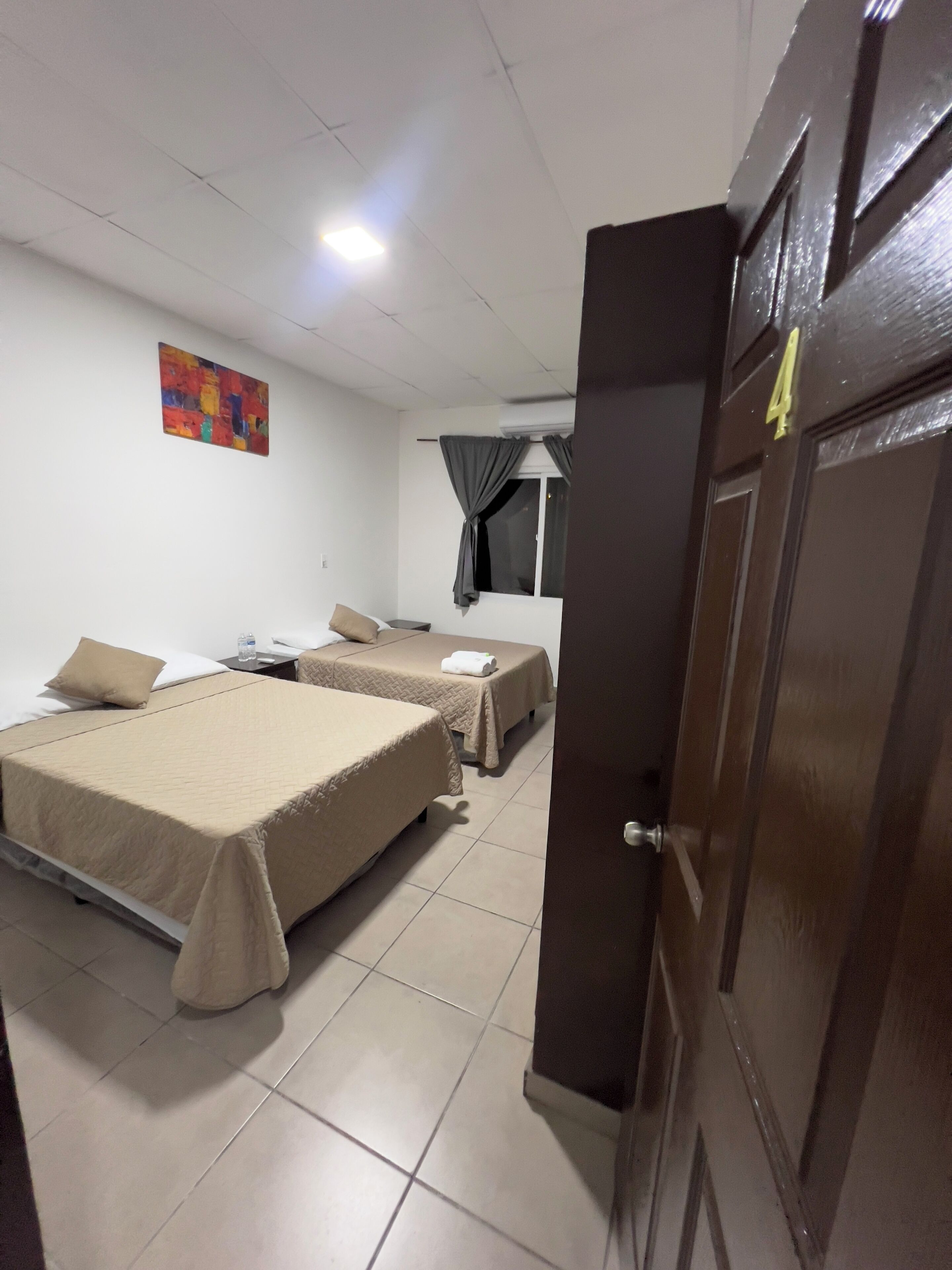 A hotel room with two beds, a small table, a wall art piece, a window with curtains, and a door with a number.