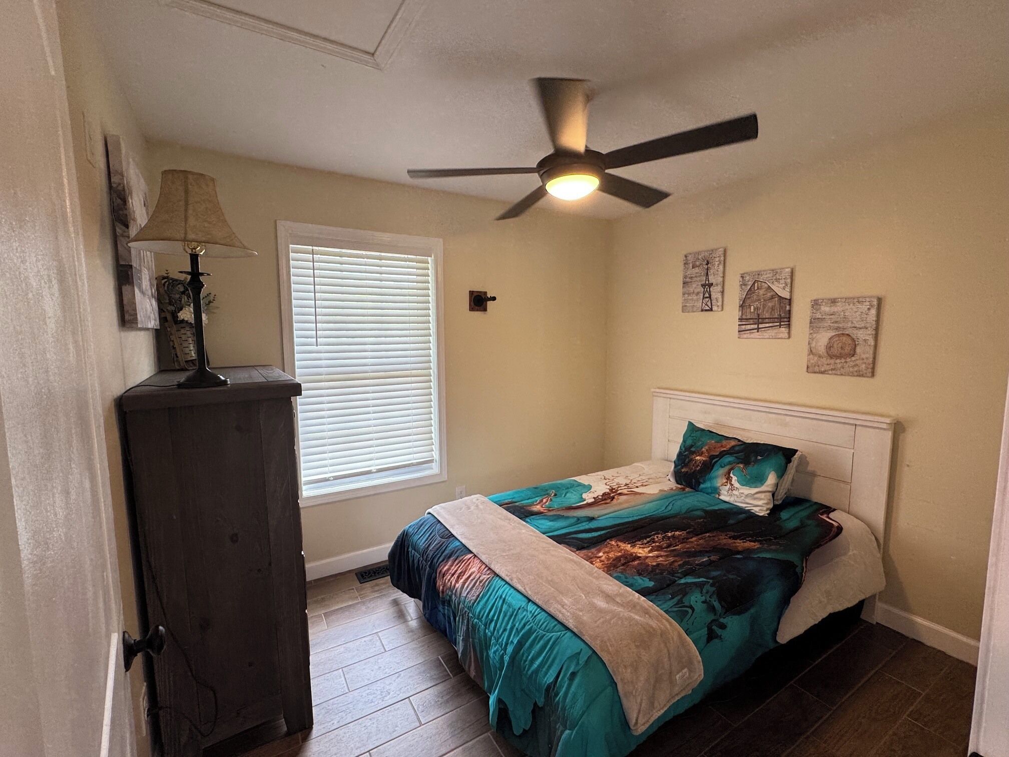 Une chambre à coucher comprenant un lit, un ventilateur de plafond, une fenêtre avec des stores, une commode et des œuvres d’art murales.