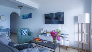 A modern living room with a sofa, armchair, coffee table, flat-screen TV, and a white desk with a lamp.