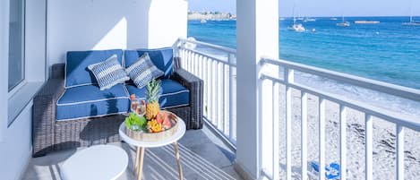 Un balcon avec un canapé en osier, une petite table ronde sur laquelle sont disposés des fruits, et une vue sur la mer et les bateaux.