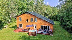 A wooden cabin with a hot tub, patio furniture, and a covered outdoor seating area surrounded by lush greenery. - Spacious 4BR / 5 Bath Home - Hot Tub / Pool Table / Ample Private Outdoor Space! (Pittsfield)