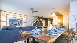 Un espace repas moderne avec une table en bois disposée pour six personnes, entourée de chaises rembourrées. Un canapé bleu est placé à l’arrière-plan, et la pièce est agrémentée d’un ventilateur de plafond, de horloges murales et d’une grande fenêtre.