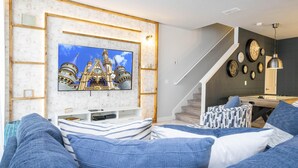 Un salon moderne doté d’un grand téléviseur à écran plat fixé sur un mur en briques blanches, d’un escalier menant à un niveau supérieur et d’un coin repas avec une table de billard.