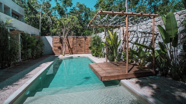 A swimming pool with a wooden deck and a pergola, surrounded by lush greenery and a stone wall. - Jungle studio (Tulum)