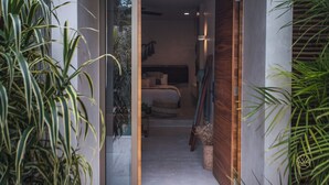 A narrow hallway leading to a bedroom with a bed, a bedside table, and a wall-mounted TV. There are potted plants on either side of the hallway. - Jungle studio (Tulum)