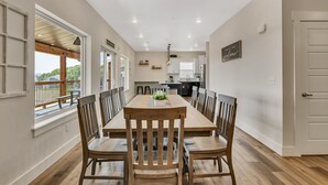 Uma sala de jantar espaçosa com uma longa mesa de madeira e cadeiras, uma cozinha ao fundo e vista para o exterior através de grandes janelas.