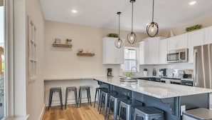 Cozinha moderna com armários brancos, uma ilha grande com banquetas, eletrodomésticos de aço inoxidável e iluminação pendente.