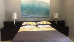 Un dormitorio con cama, dos lámparas de noche, un cuadro grande de una ola en la pared y una mesita de noche de color oscuro.