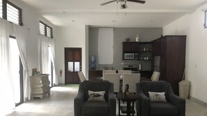 Un salón amplio con dos sillones, una mesa de centro y un comedor con mesa y sillas. Se ve una cocina con microondas y estufa al fondo.