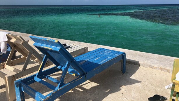 Vista a la playa con una terraza de madera, sillas reclinables azules y una playa de arena.
