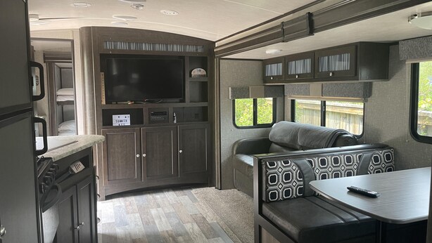 Veloplyst autocamper indvendigt med et køkkenområde, et siddeområde med et bord og et stort TV ophængt over køkkenet.