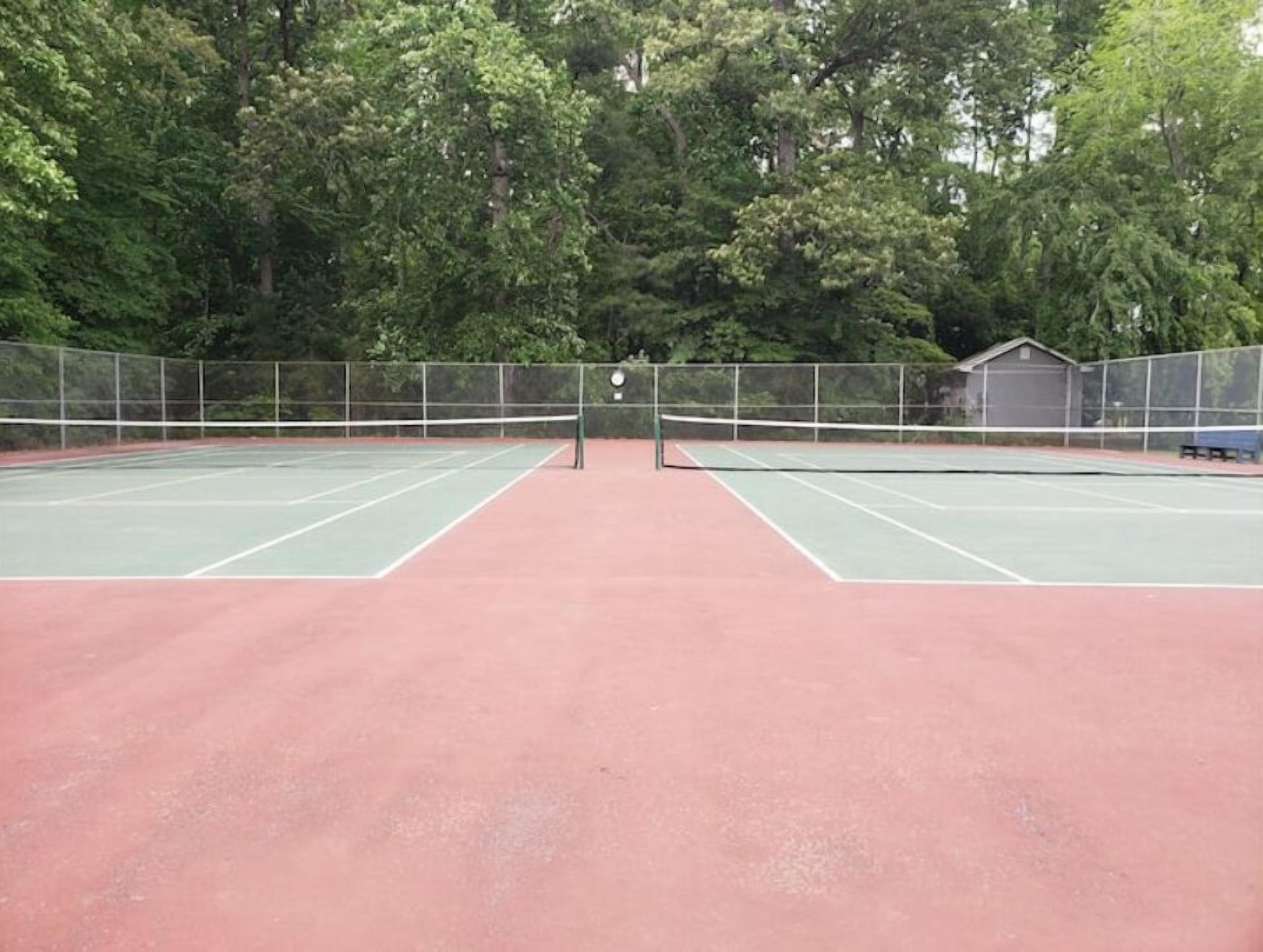An outdoor tennis court with a net in the center, surrounded by a fence and trees.
