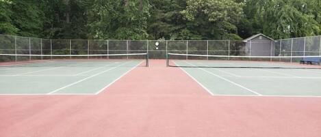 An outdoor tennis court with a net in the center, surrounded by a fence and trees.