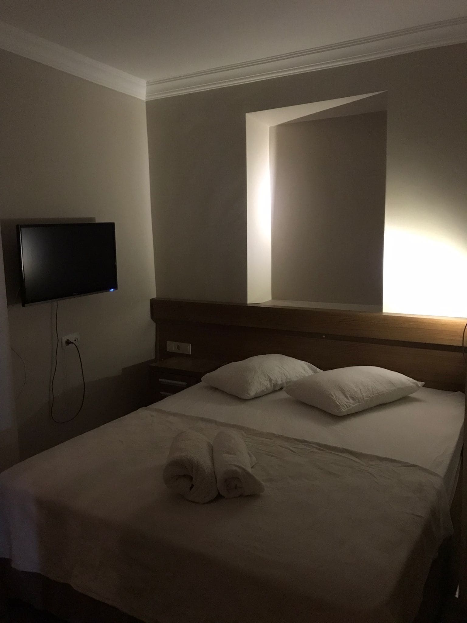 Quarto de hotel com cama, televisão de tela plana na parede, mesa de cabeceira e criado-mudo.