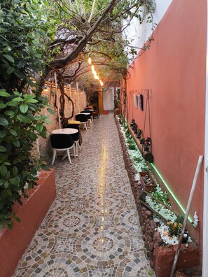 A narrow outdoor corridor with tiled flooring, tables, chairs, and a hanging light fixture. - Lavinya Pansiyon (Kusadasi)