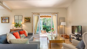 A living room with a sofa, armchairs, a coffee table, a TV, and a view of the garden through the door. - 3 bed + villa near Gordes with air-con, pool and stunning views over the Luberon (Cabrières-d'Avignon)