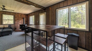 Een rustiek blokhutinterieur met een grote houten tafel, bankjes, een open haard en grote ramen met uitzicht op een bos.