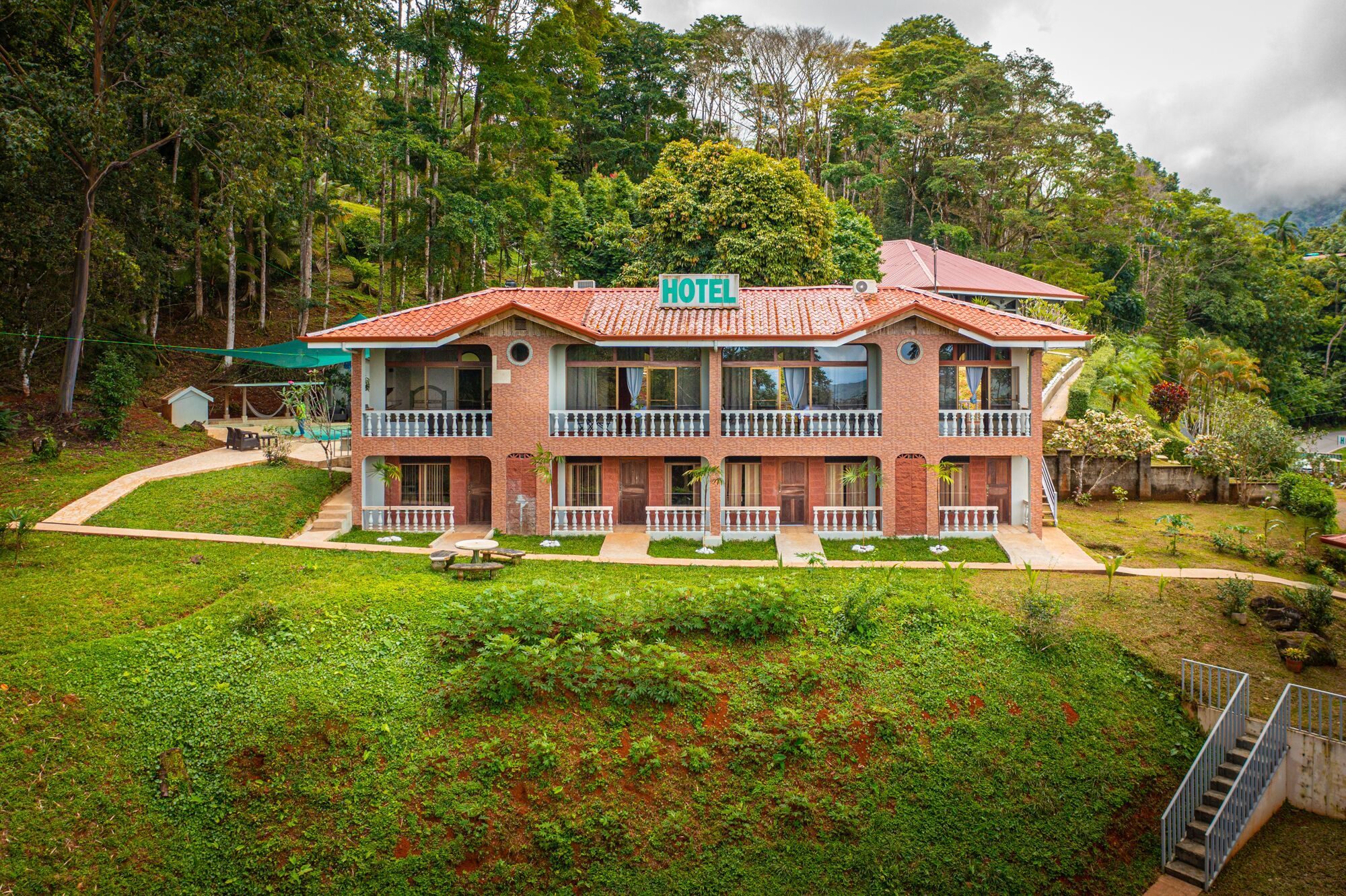 Un hotel de dos pisos con tejado de tejas rojas, varios balcones y un exuberante entorno de jardín.