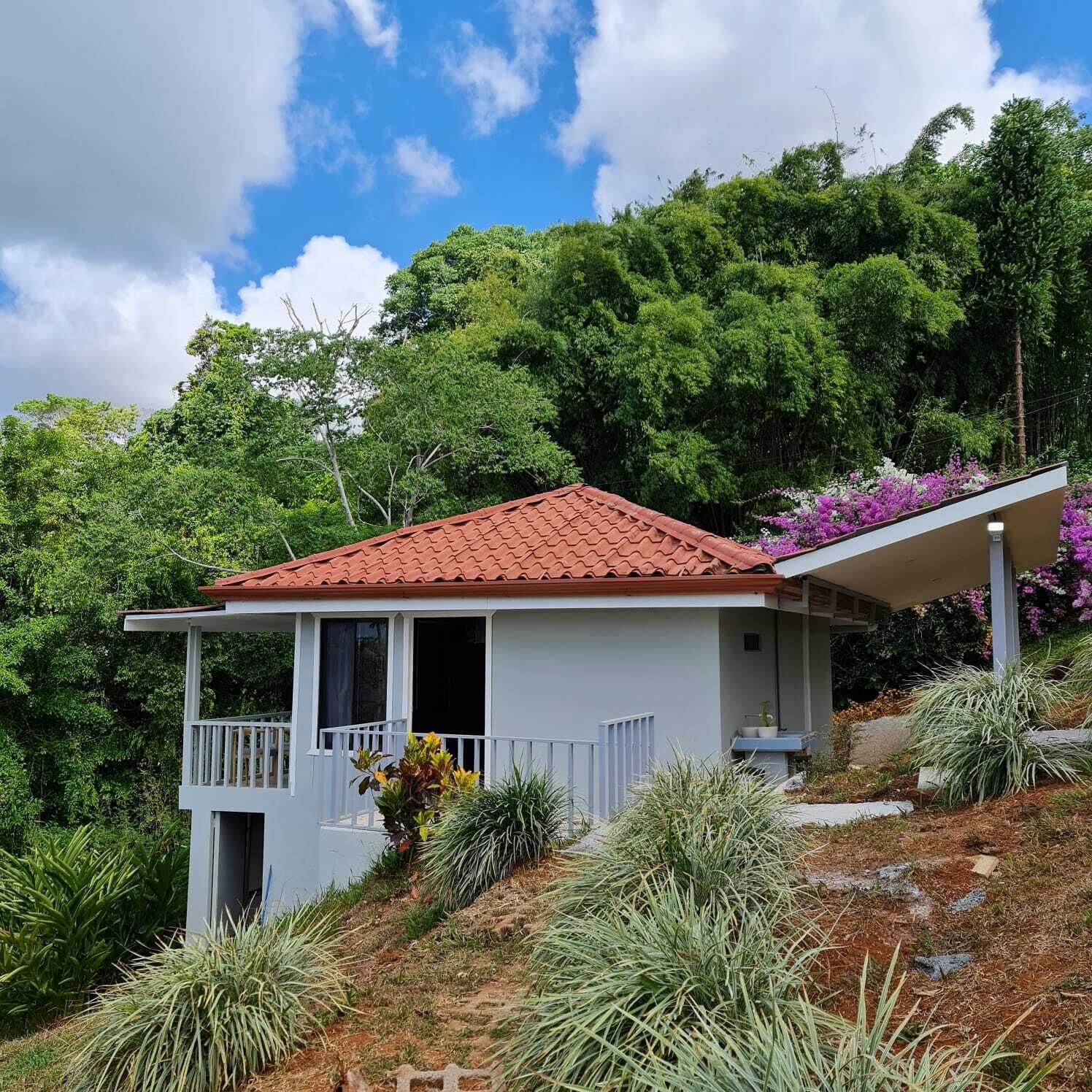 A single-story house with a red-tiled roof, a balcony, and a garden with various plants and flowers.