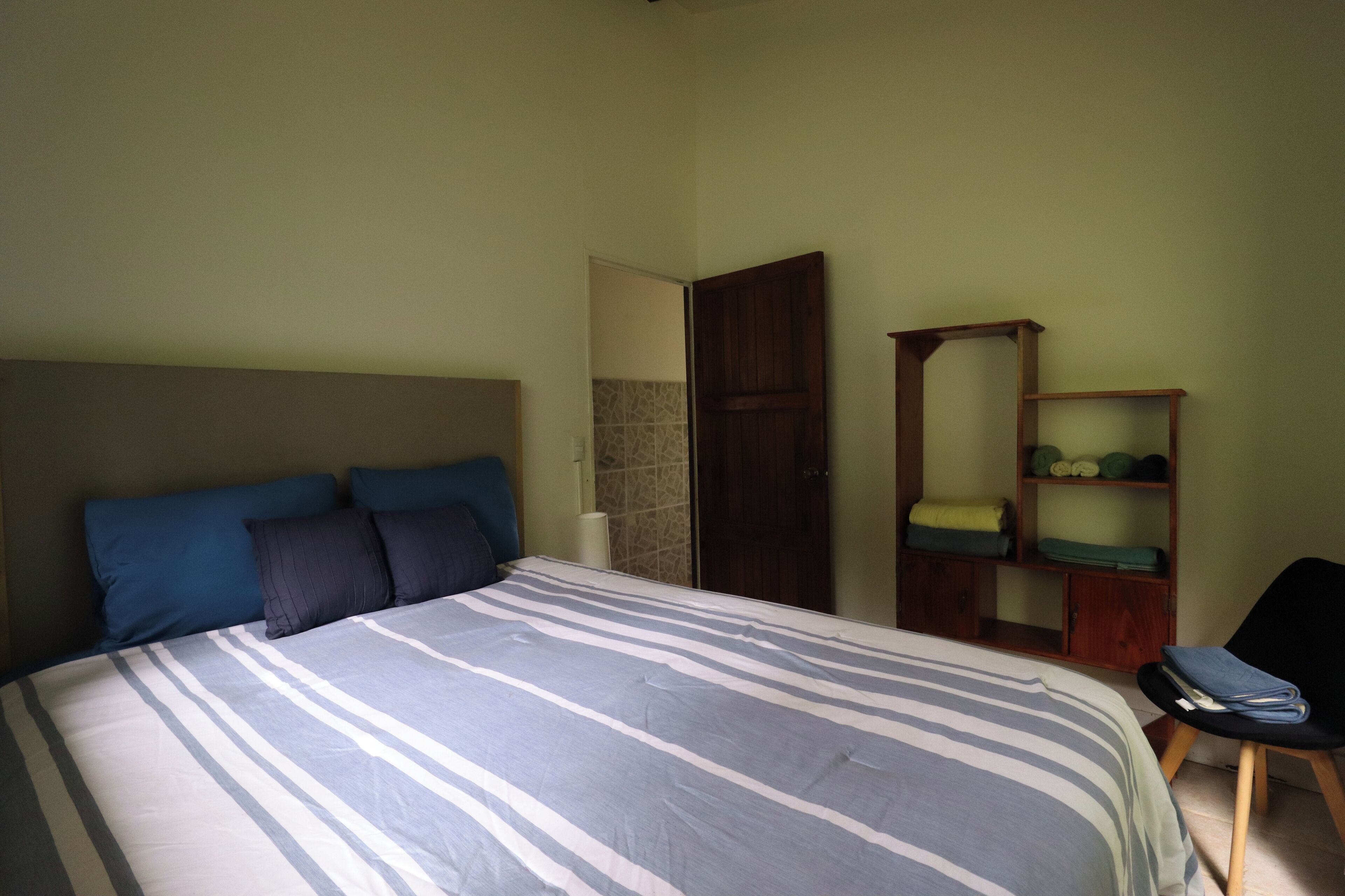 A bedroom with a bed, pillows, a chair, and a wooden shelf with folded clothes.