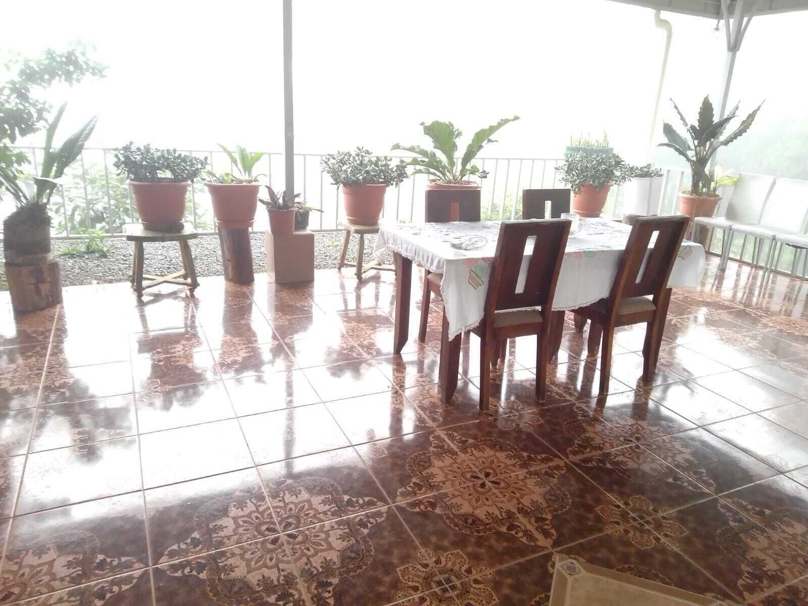 Een overdekt terras met betegelde vloer, een eettafel en plantenpotten langs de rand.