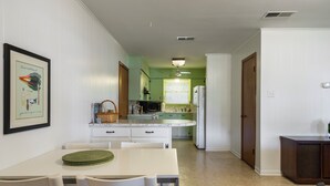 Une cuisine avec des meubles blancs, une table à manger avec des chaises, un réfrigérateur et une fenêtre avec des stores.