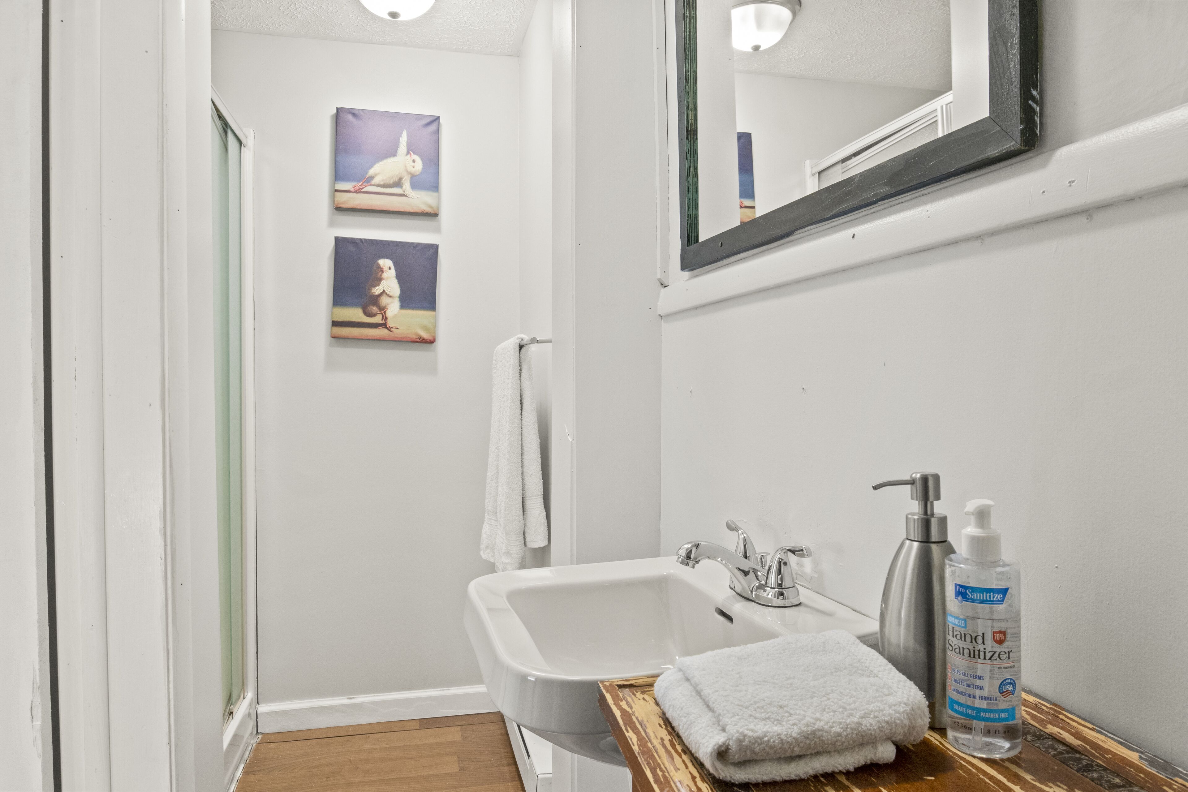 Un baño con lavamanos blanco, tocador de madera, botella de gel antibacterial y espejo.