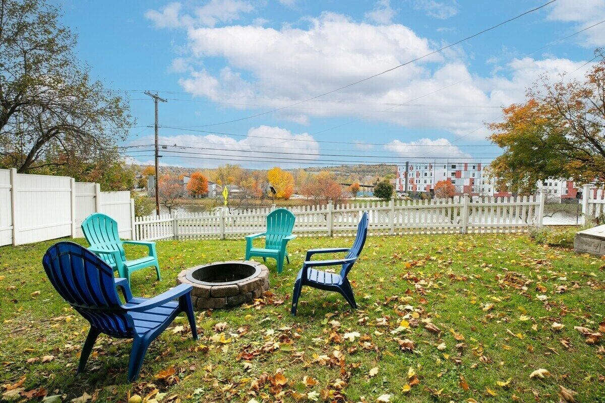 A backyard with three Adirondack chairs, a fire pit, and a white picket fence.