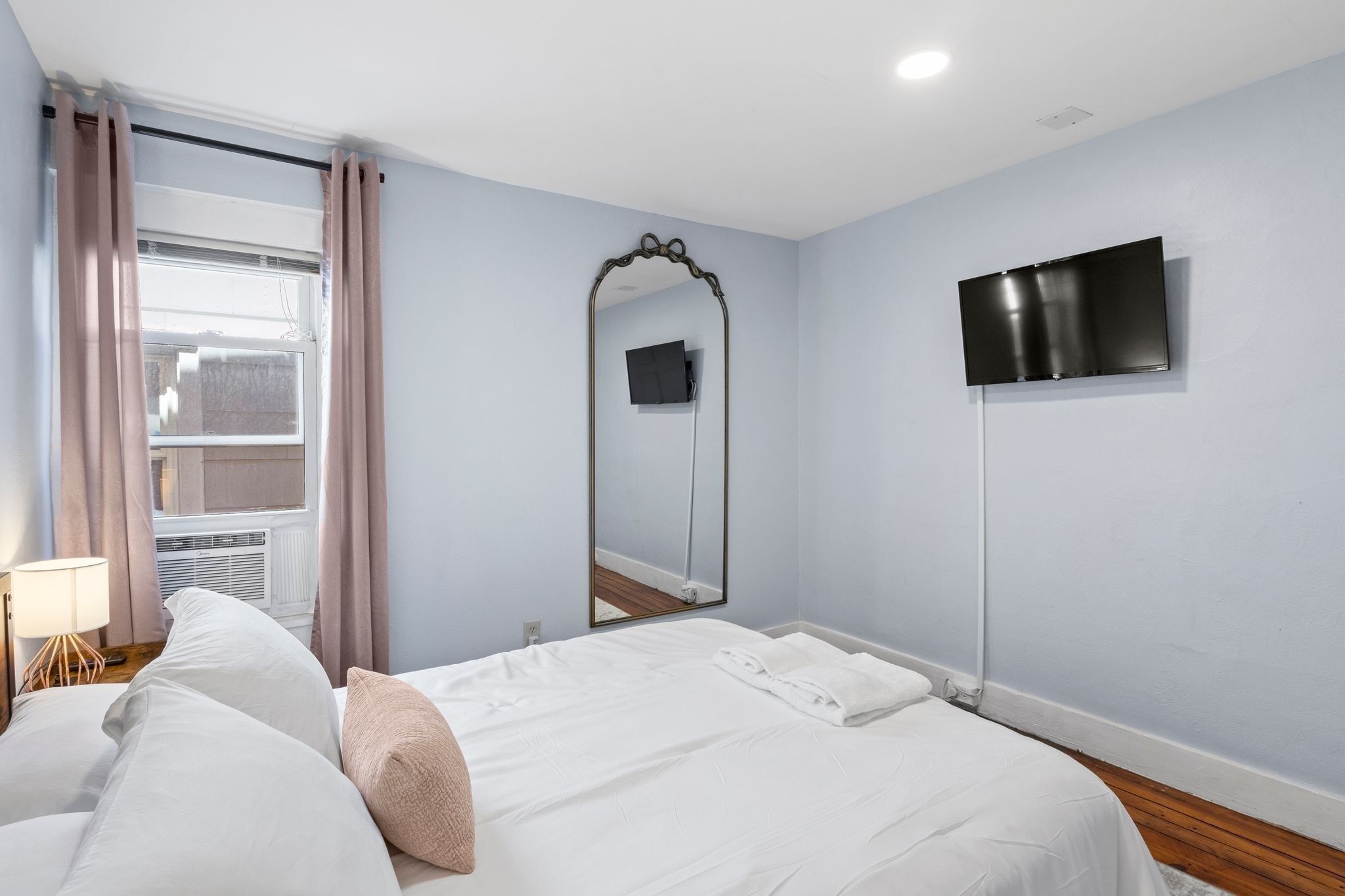 A hotel room with a bed, a TV mounted on the wall, a window with curtains, and a mirror.