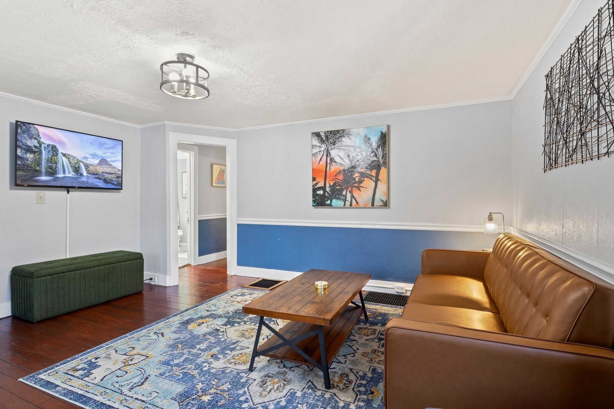 A living room with a brown leather sofa, a wooden coffee table, a green bench, a blue patterned rug, a TV on the wall, and two paintings.