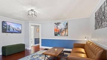 A living room with a brown leather sofa, a wooden coffee table, a green bench, a blue patterned rug, a TV on the wall, and two paintings.